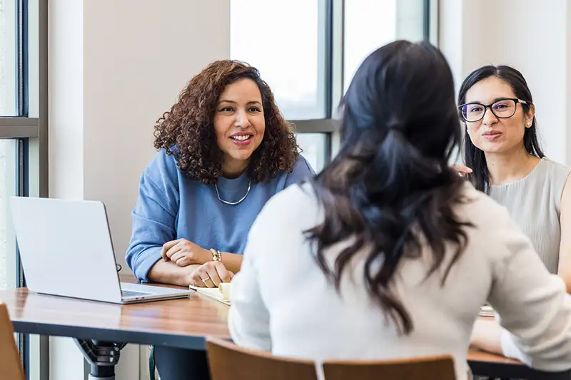 Three women at a desk with notepads and laptops talking to eachother